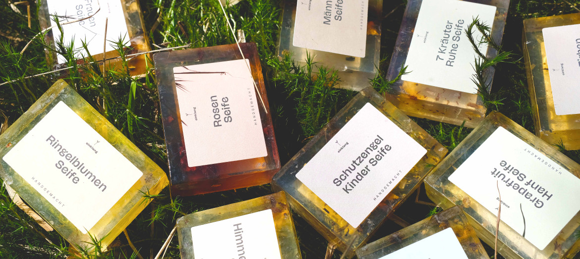 Mehrere handgemachte Glycerinseifen von Himmelgrün in unterschiedlichen Sorten wie Rosen-, Ringelblumen- und Kräuterseife, dekorativ im Gras liegend und von Natur umgeben.