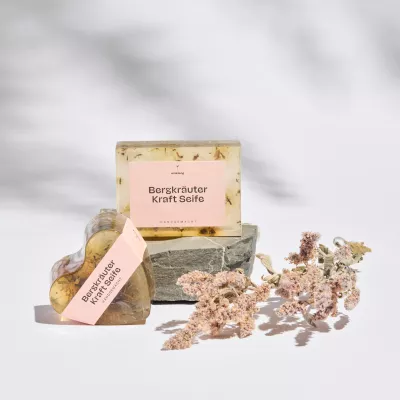 Handgemachte Bergkräuter Kraft Seife von Himmelgrün als transparente Glycerinseife mit Kräutern, präsentiert auf einem Naturstein neben getrockneten Blüten und einer herzförmigen Seife.