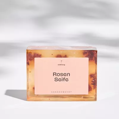 Transparente, handgemachte Rosenseife von Himmelgrün mit eingearbeiteten Rosenblüten, rechteckig geformt und mit schlichtem Etikett auf hellem Hintergrund präsentiert.